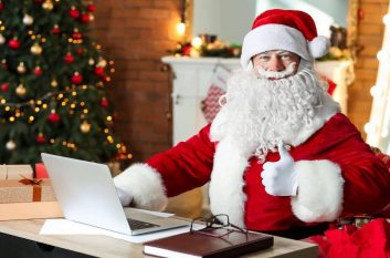 Santa's going over his nice and naughty cybersecurity lists. He sits at a laptop and gives a thumbs up to the camera. There's a Christmas tree behind him.
